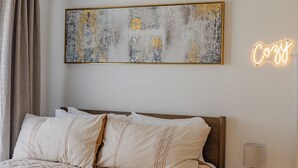 Ein ordentlich bezogenes Bett mit Kopfteil, ein an der Wand befestigtes abstraktes Gemälde und ein leuchtendes Neon-Schild mit der Aufschrift 'Cozy'.