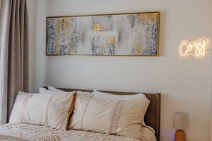 A neatly made bed with a headboard, a wall-mounted abstract painting, and a neon sign that reads "Cozy". - Modern 1BR w/ Massive Deck & City Views (Nashville)