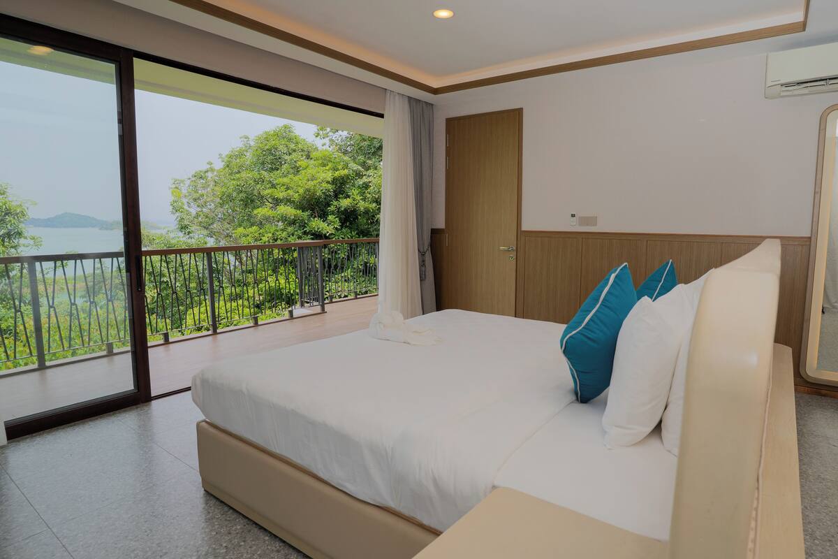 A modern bedroom with a large bed, a wooden headboard, and a balcony with a view of greenery and water.
