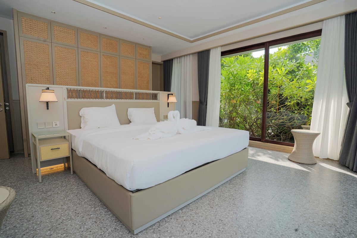 A modern hotel room with a large bed, a nightstand, a lamp, and a view of greenery through the window.