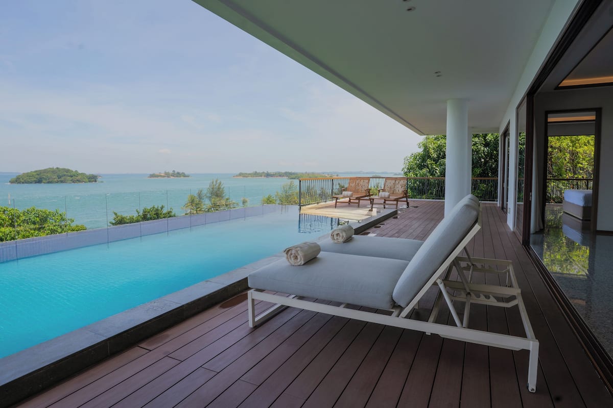 A wooden deck with a pool, two lounge chairs, and a view of the sea and islands.