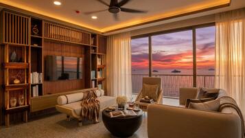 A modern living room with a large TV, bookshelves, a sofa, and a view of the sunset over the water.