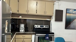 Eine kleine Küche mit beigen Schränken, einem Edelstahlkühlschrank, einer Kaffeemaschine, einem Backofen und einem Glastisch mit blauen Stühlen.