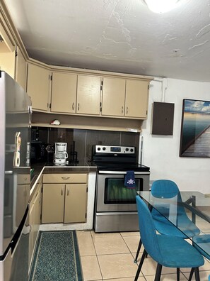 A compact kitchen with beige cabinets, a stainless steel refrigerator, a coffee maker, an oven, and a glass table with blue chairs. - Charming 1-bedroom apartment with AC in central Hagåtña (Hagåtña)