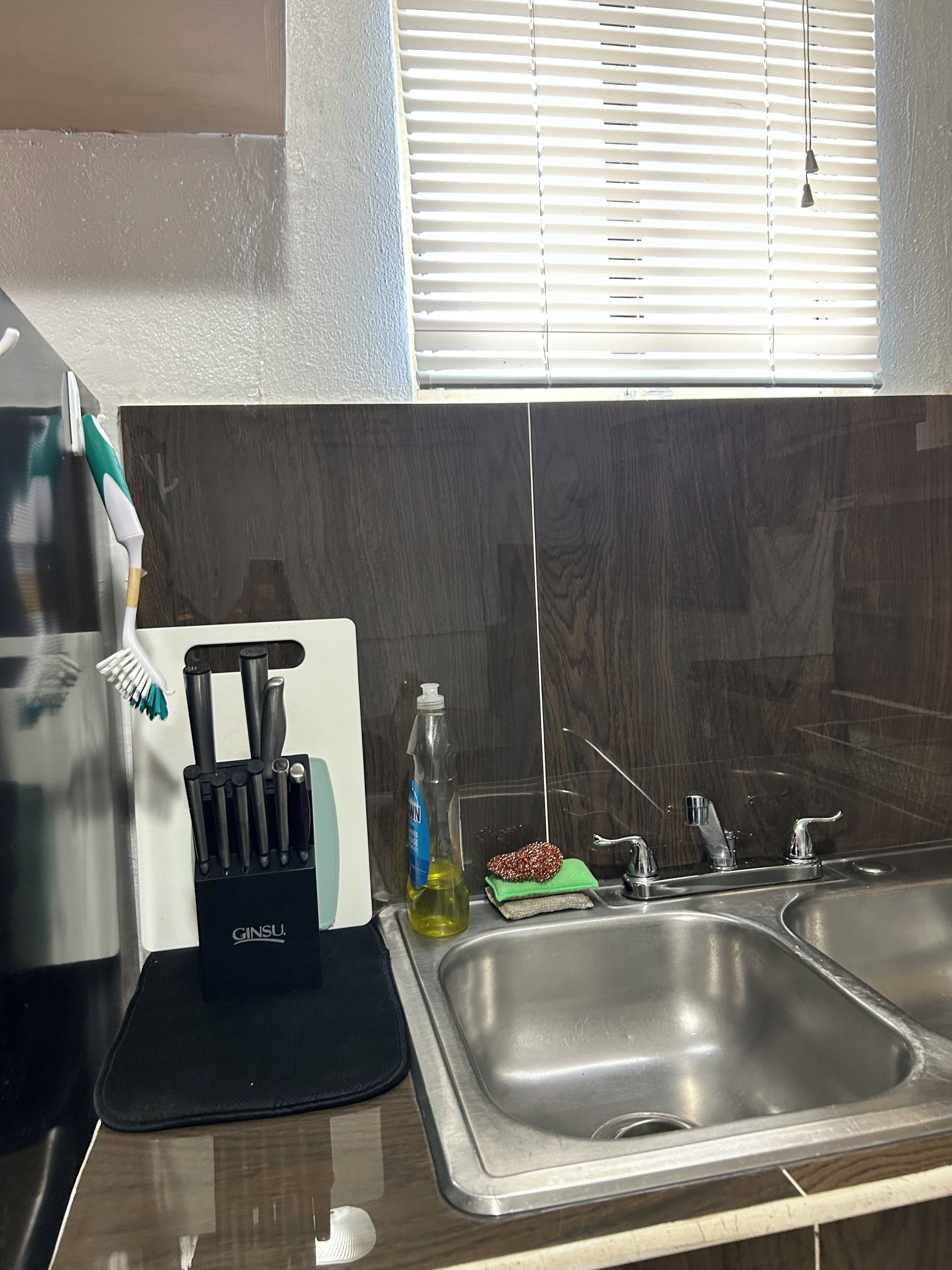 A kitchen with a stainless steel sink, a knife block, a bottle of dish soap, and a green cloth.