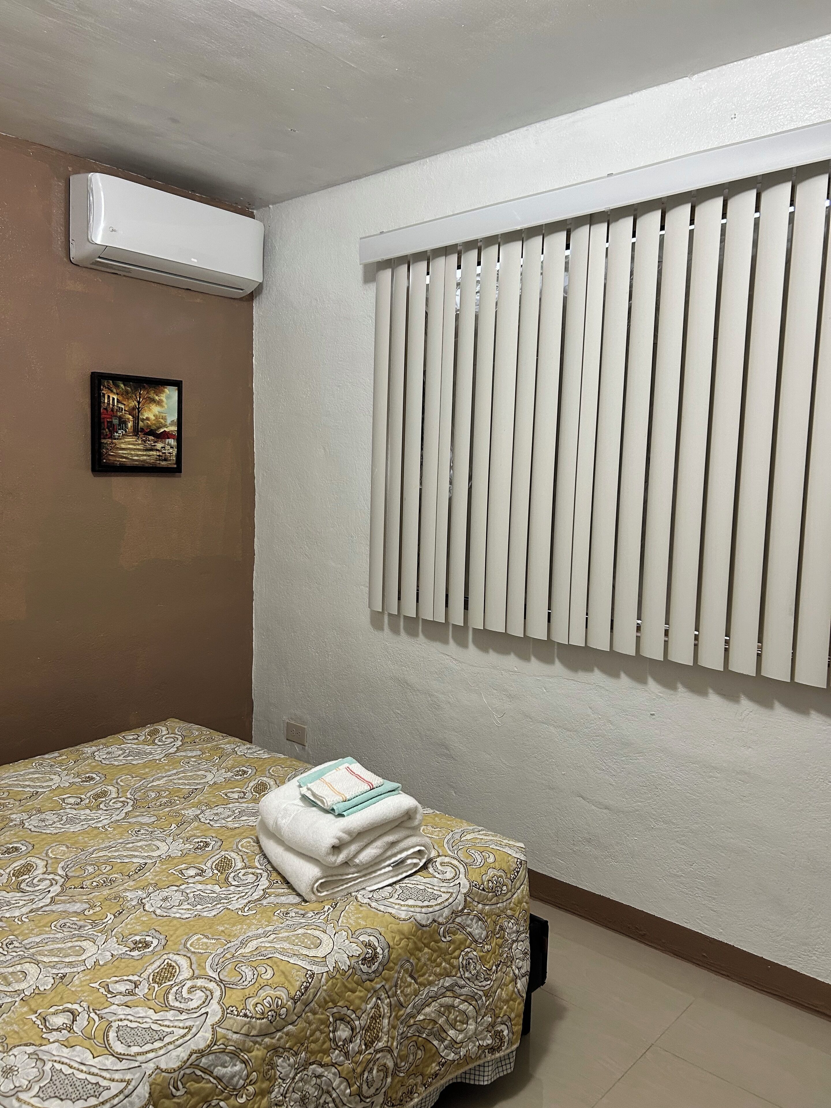 Une chambre avec un lit, un climatiseur, une fenêtre avec des stores et un tableau au mur.