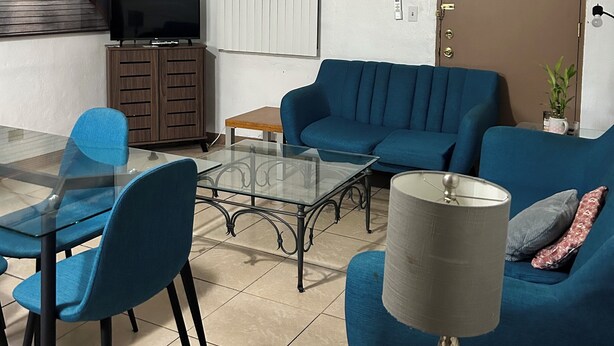 Ein Wohnzimmer mit Glastisch, blauen Stühlen, einem blauen Sofa, einer Lampe und einem Fernseher.
