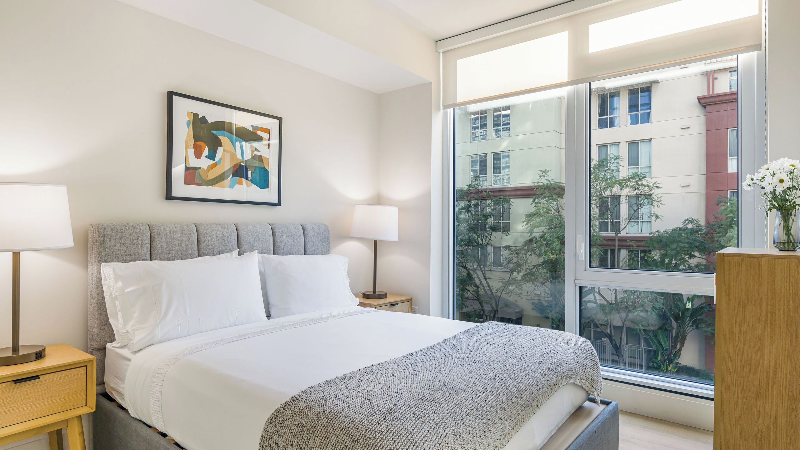A modern bedroom with a bed, bedside lamps, a framed abstract painting, and a large window with a view of buildings and trees.