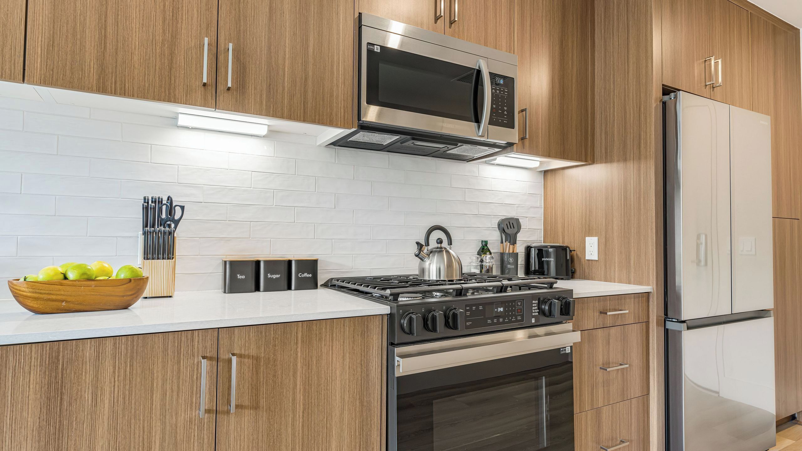 A modern kitchen with wooden cabinets, a stainless steel stove, a microwave, and a refrigerator.