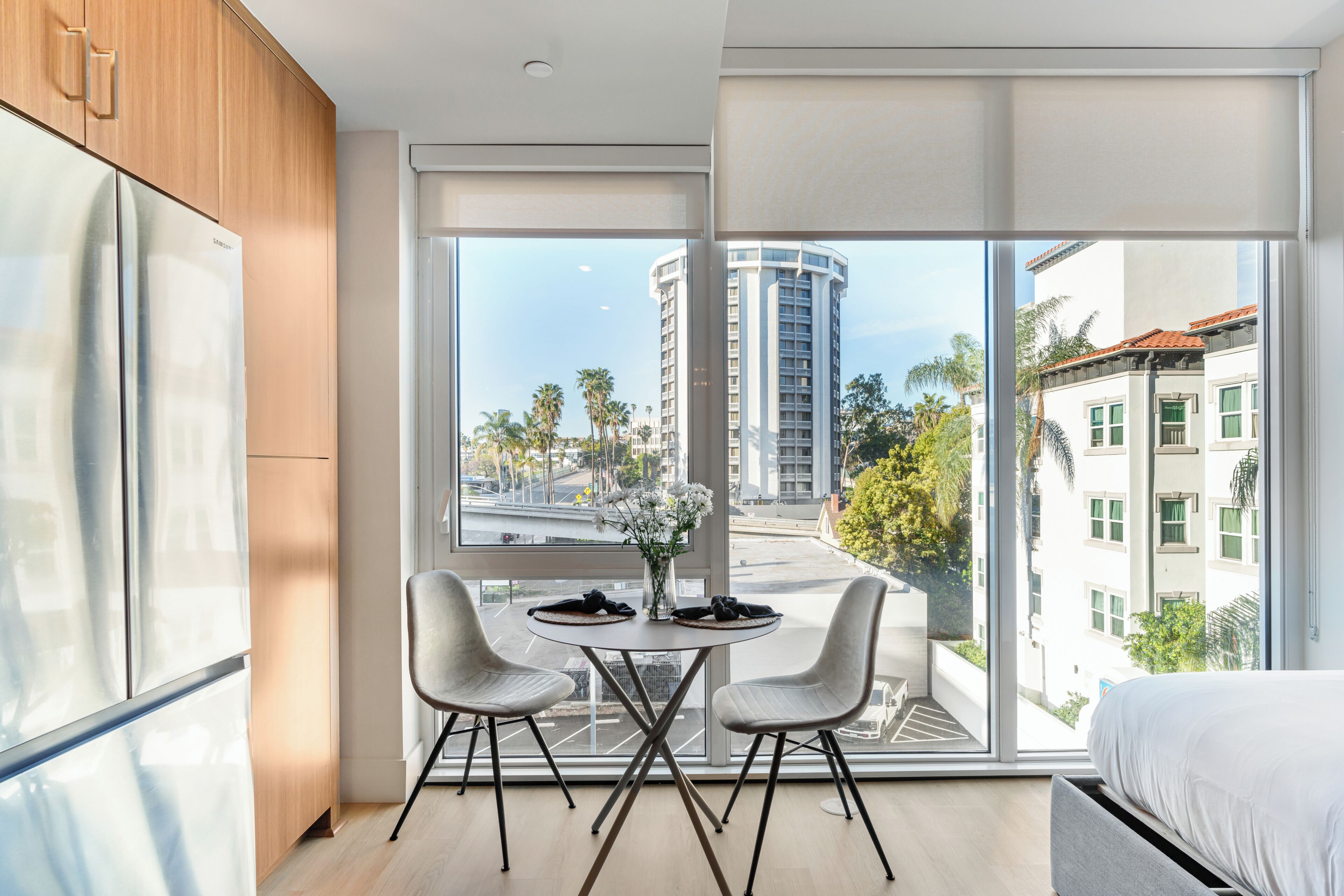 A modern hotel room with a large window, a dining table with chairs, a bed, and a refrigerator.