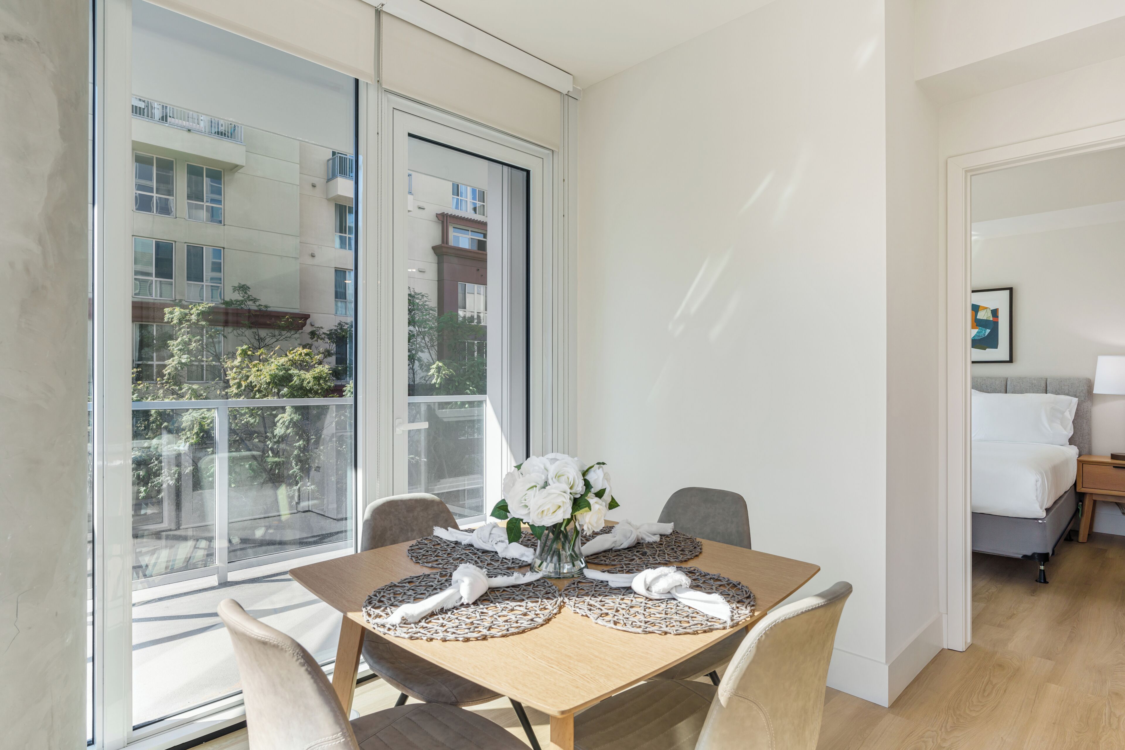 A modern dining area with a large window, a round table with placemats, and a vase with flowers.