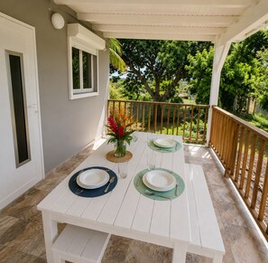 A white wooden table set for four with a vase of flowers, on a balcony with a view of trees and a wooden railing. - Canopée Créole | House with a garden and a hot tub (Petit-Canal)