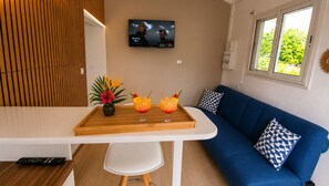 A modern living room with a blue sofa, a wooden coffee table with drinks, a TV on the wall, and a window with a view. - Canopée Créole | House with a Garden and Hot Tub (Petit-Canal)