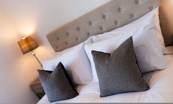 Una cama impecable con un cabecero capitonado, varias almohadas y una lámpara fijada a la pared.