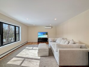 Un salón amplio con un ventanal grande, un televisor de pantalla plana, un sofá gris y una alfombra estampada.