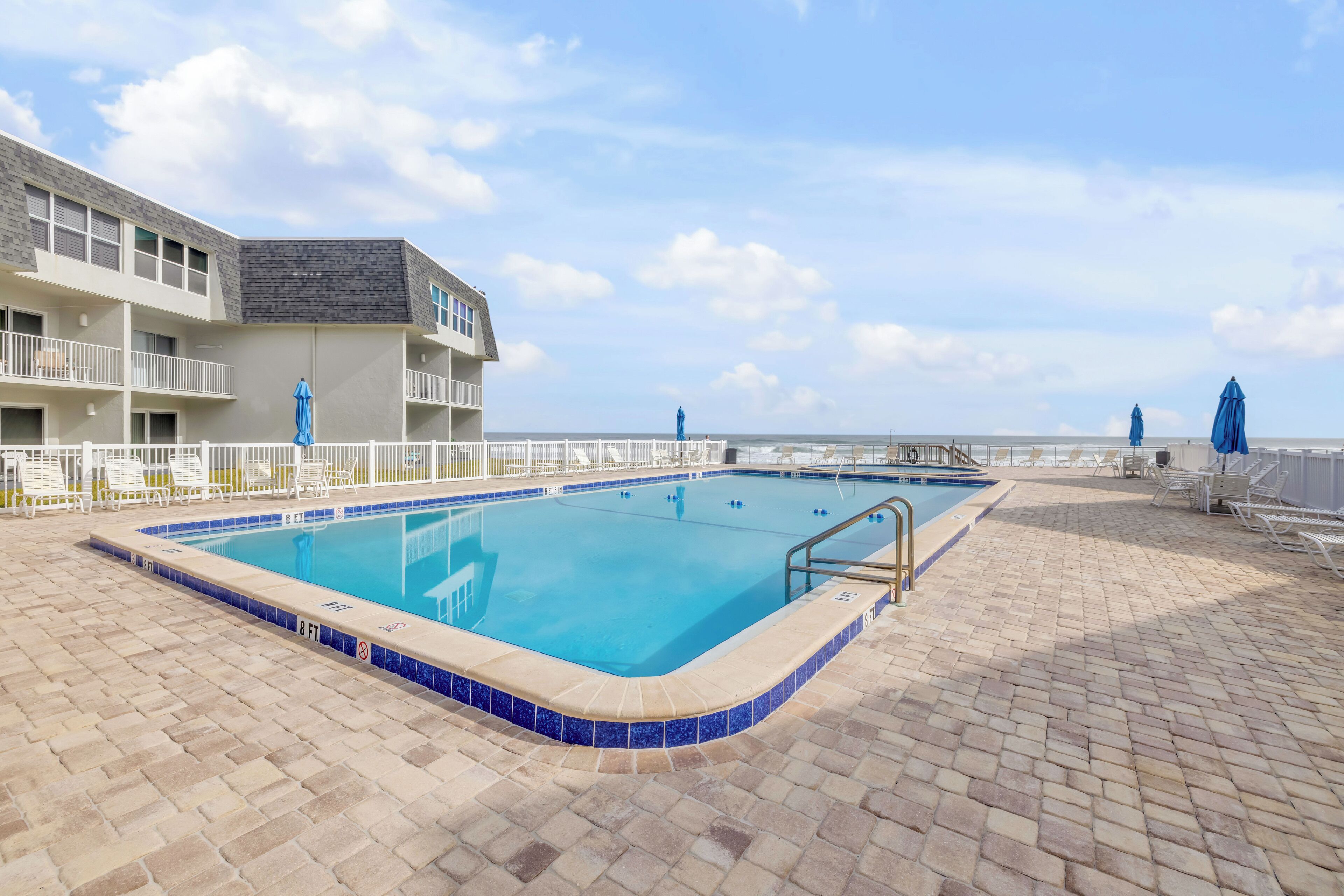 Une piscine dont le bord est recouvert de carreaux, une clôture blanche et, au fond, un bâtiment doté de balcons.