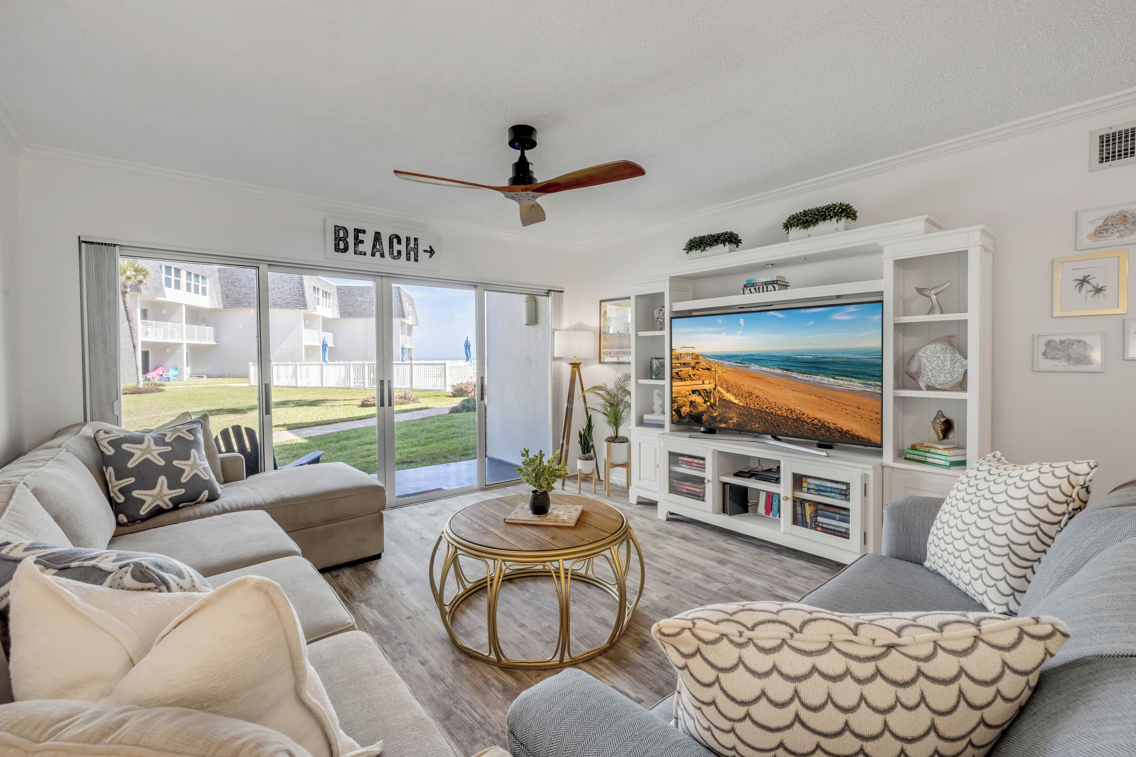 A living room with a large TV, a ceiling fan, and a view of the beach through the sliding glass doors.
