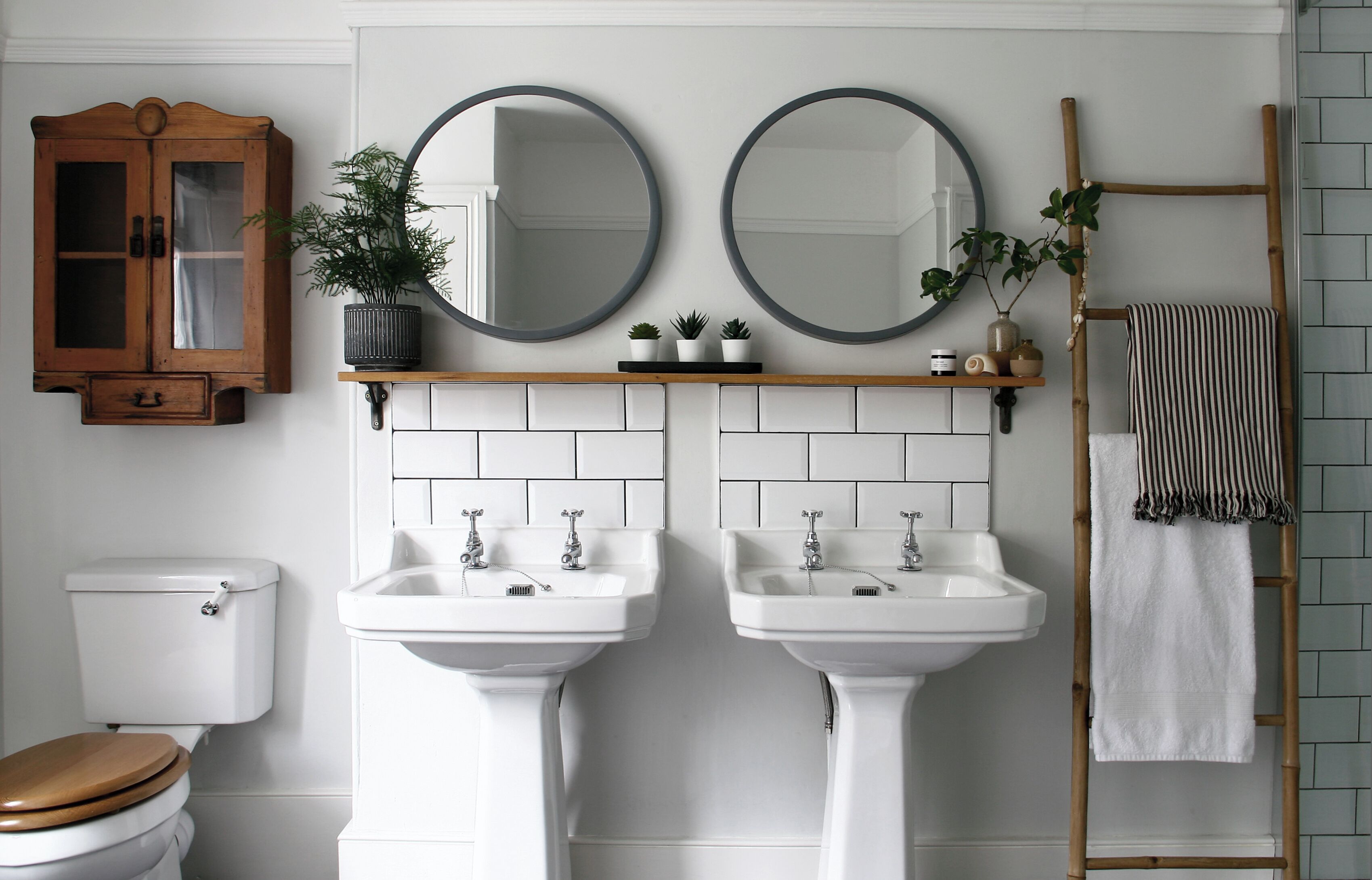 A bathroom with two sinks, a toilet, a wooden cabinet, and two round mirrors.