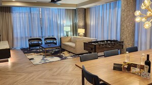 Une chambre d’hôtel avec une table à manger en bois, deux canapés, un baby-foot, un lustre et une grande fenêtre avec des rideaux.