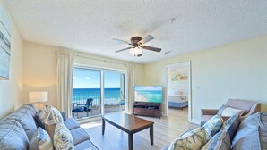 A spacious living room with a large window overlooking the ocean, a ceiling fan, and a television. - Beautiful Beachfront 3BR Condo with Pool! FREE Summer Beach Service! (Destin)