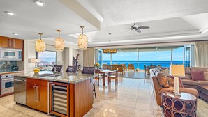 Een ruime woonkamer met een groot raam met uitzicht op de oceaan, een keuken met moderne apparatuur, en een eetgedeelte met houten meubels.