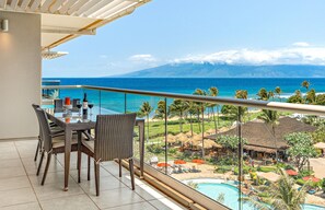 Un balcon avec une table et des chaises, donnant sur une piscine et une plage bordée de palmiers.