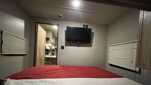 A bedroom with a red and white bedspread, a mounted TV, and a door leading to a bathroom. - Come Settle in this cozy modern style RV, Nestled in the quiet country side. (Bushnell)