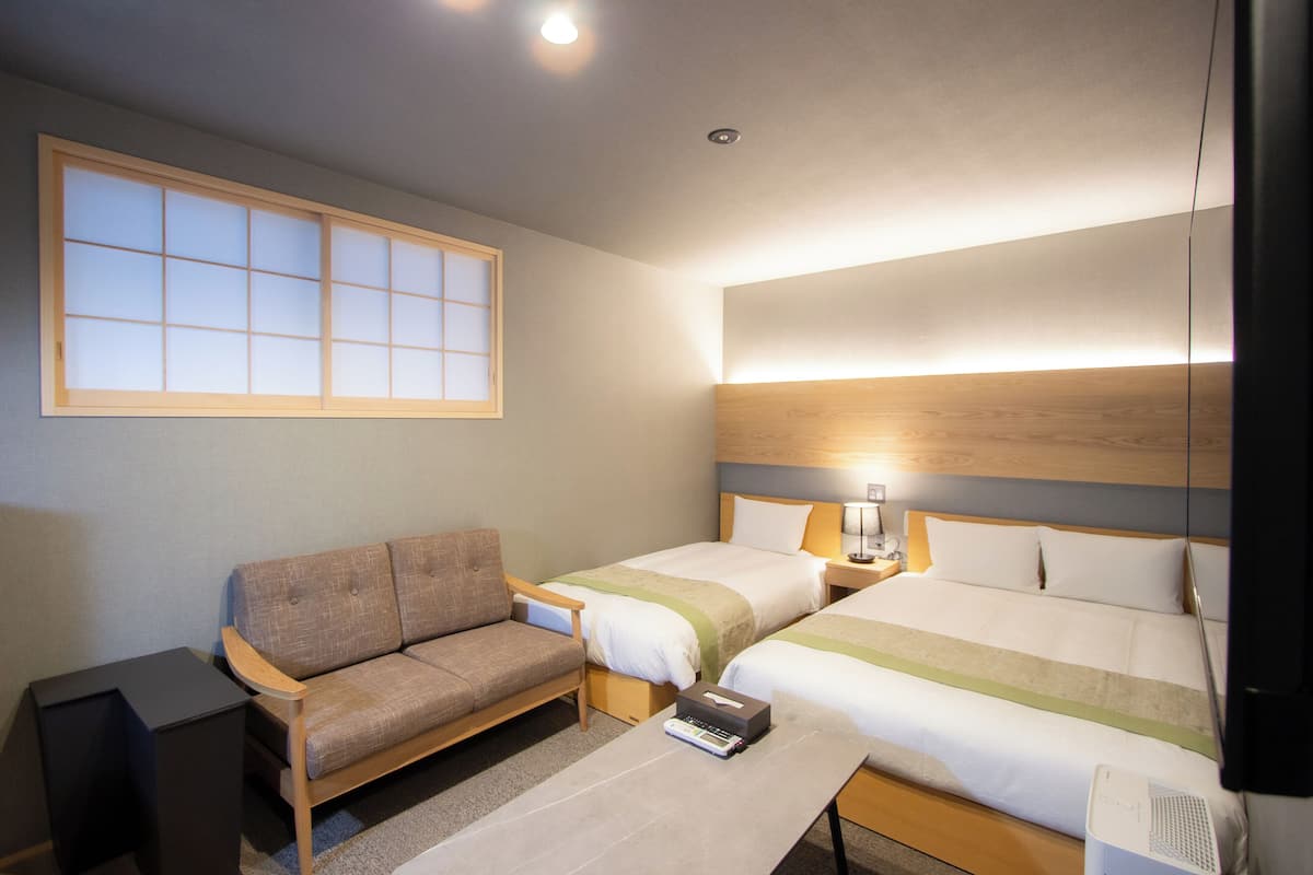 A hotel room with two beds, a sofa, a bedside table, a lamp, and a window.