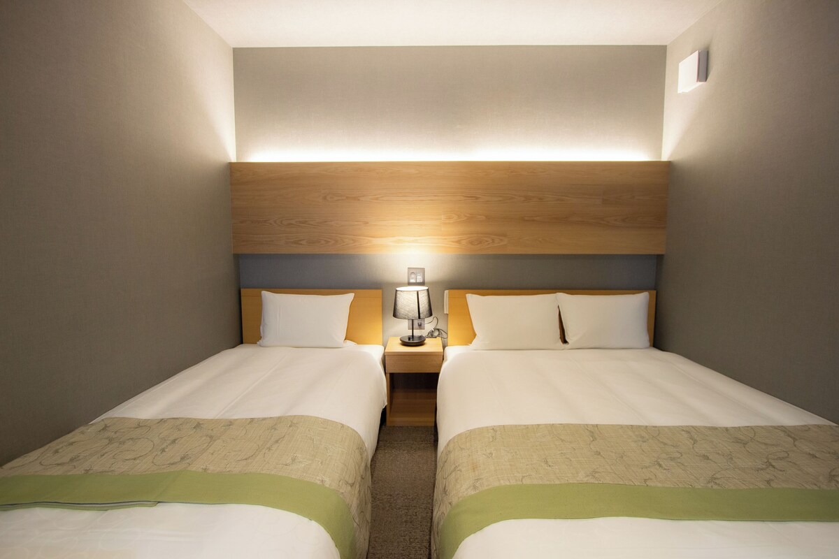 A hotel room with two beds, a wooden headboard, a nightstand with a lamp, and a carpeted floor.