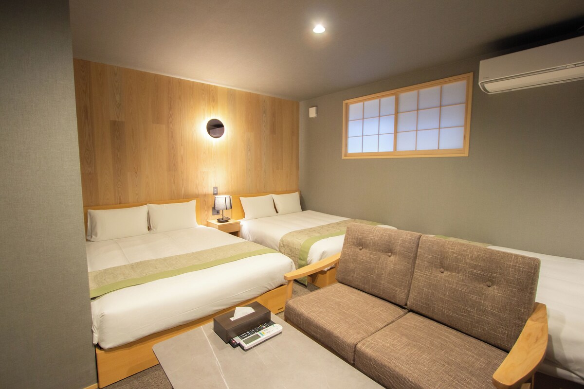 A hotel room with two beds, a sofa, a bedside table with a lamp, and a window with a view.