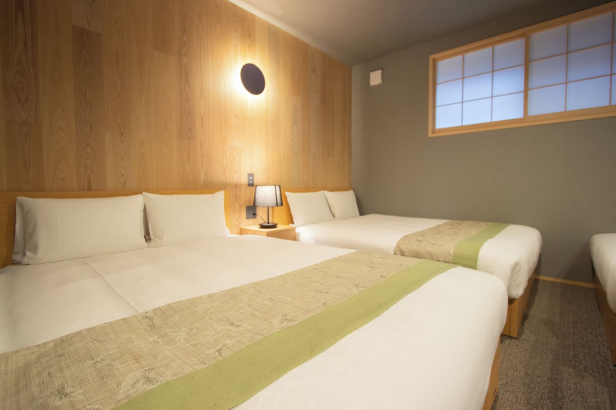 A hotel room with two beds, a nightstand, a lamp, and a window with blinds.