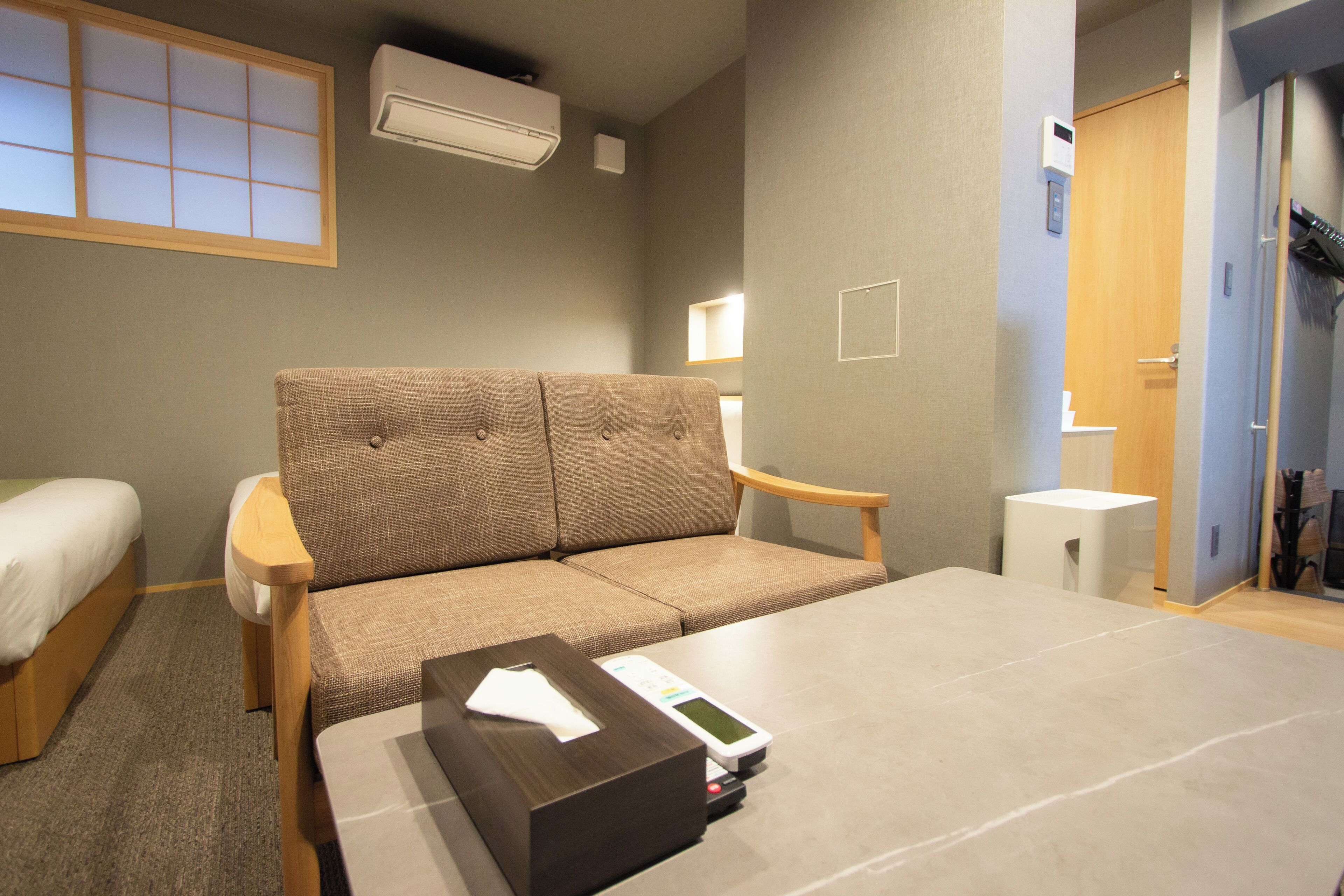 Hotellihuone, jossa on sohva, pieni pöytä, yöpöytä, ikkuna, josta on näkymä, ilmastointilaite ja ovi, joka johtaa kylpyhuoneeseen.