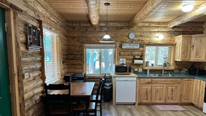 A log cabin interior with a wooden dining table, chairs, a kitchen area with a microwave, sink, and cabinets, and a clock on the wall. - Hand Crafted Log Cabin in Island Park (Island Park)