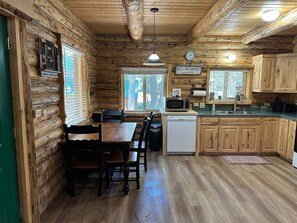 A log cabin interior with a wooden dining table, chairs, a kitchen area with a microwave, sink, and cabinets, and a clock on the wall. - Hand Crafted Log Cabin in Island Park (Island Park)