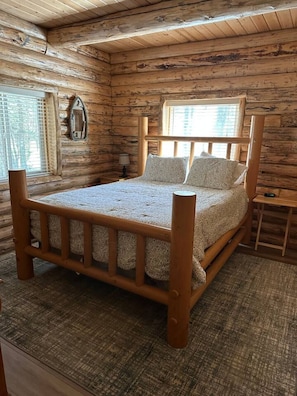 A wooden cabin bedroom with a large bed, a window with blinds, and a small bedside table. - Hand Crafted Log Cabin in Island Park (Island Park)
