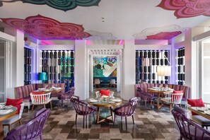 Un comedor con mesas redondas, sillas moradas y un mural colorido de fondo.