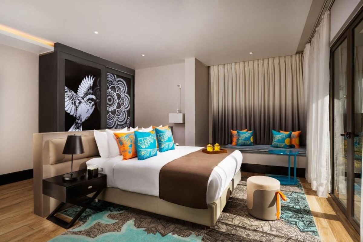 A modern hotel room with a large bed, a bedside table, a lamp, a wall art piece, a window with curtains, and a patterned rug.