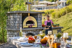 An outdoor pizza oven with a stone facade, a chef preparing food, and a table set with plates, glasses, and a bottle. - Norbu The Montanna, Dharamshala - Ihcl Seleqtions (Dharamshala)