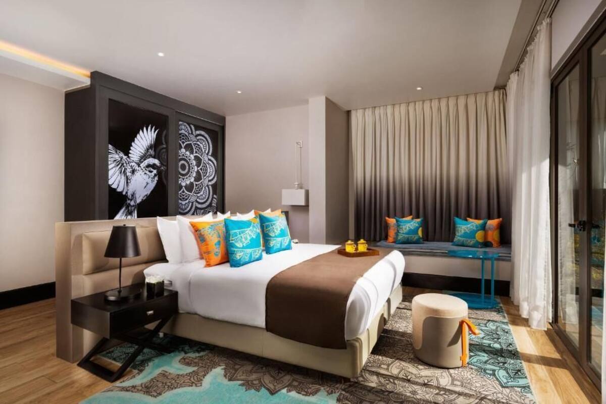 Ein modernes Hotelzimmer mit einem großen Bett, einem Nachttisch, einer Lampe, einem Wandbild, einem Fenster mit Vorhängen und einem gemusterten Teppich.