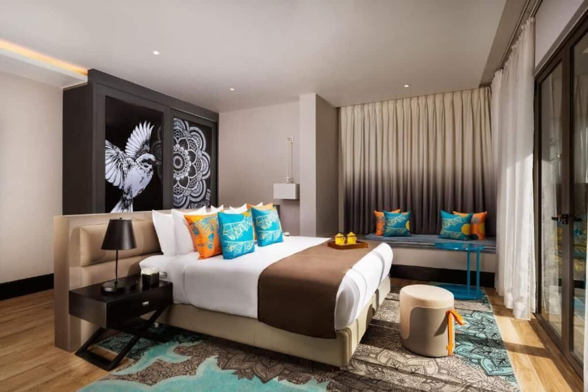 A modern hotel room with a large bed, a bedside table, a lamp, a wall art piece, a window with curtains, and a patterned rug.