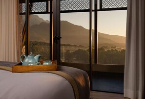 Un dormitorio con un ventanal grande, una cama, una bandeja de madera con una tetera y tazas, y vistas a montañas.