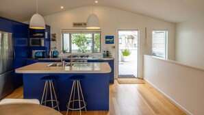 A modern kitchen with blue cabinets, a central island with stools, stainless steel appliances, and pendant lighting. - Beachfront 25 The Strand | Moana (Waiheke Island)