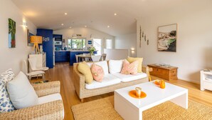 A modern living room with a sofa, coffee table, and a kitchen in the background. - Beachfront 25 The Strand | Moana (Waiheke Island)
