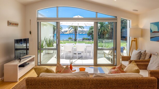 A living room with a large sliding glass door leading to an outdoor patio with a table and chairs, a television, and a sofa with cushions. - Beachfront 25 The Strand | Moana (Waiheke Island)