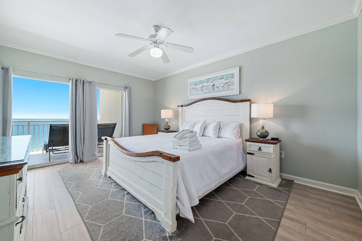 Een hotelkamer met een bed, twee nachtkastjes, een plafondventilator, een schilderij aan de muur en uitzicht op de oceaan door het raam.