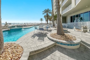 A pool area with lounge chairs and a hot tub, surrounded by palm trees and a building with balconies. - Admirals Quarters 1504 | Gulf-Front + Hot Tub (Orange Beach)