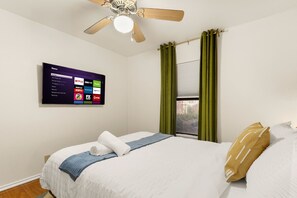 Een hotelkamer met een bed, een plafondventilator, een aan de muur bevestigde televisie en een raam met gordijnen.