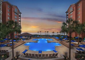 A resort pool area with multiple pools, lounge chairs, and umbrellas, surrounded by palm trees and hotel buildings. - Marriott's OceanWatch | Oceanfront 2 BR Villa (Myrtle Beach)