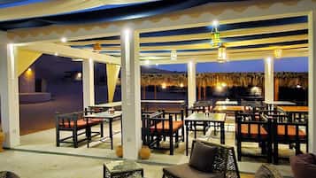 An outdoor seating area with tables and chairs, a view of the sea, and thatched umbrellas.