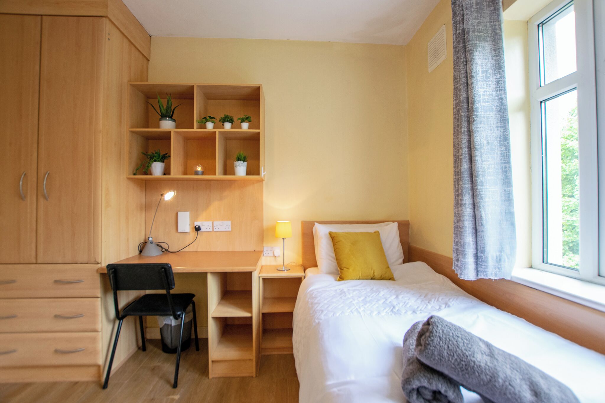A bedroom with a bed, a desk, a chair, and shelves with plants.